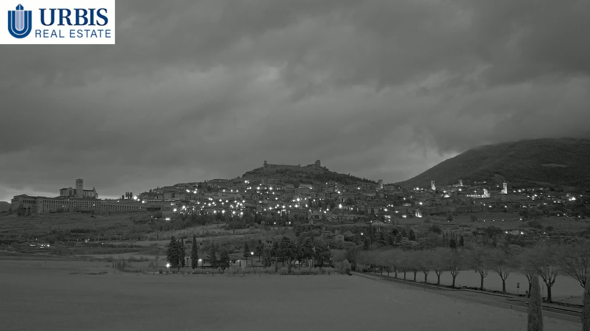 A hilltop town is illuminated at night under a dark, cloudy sky.