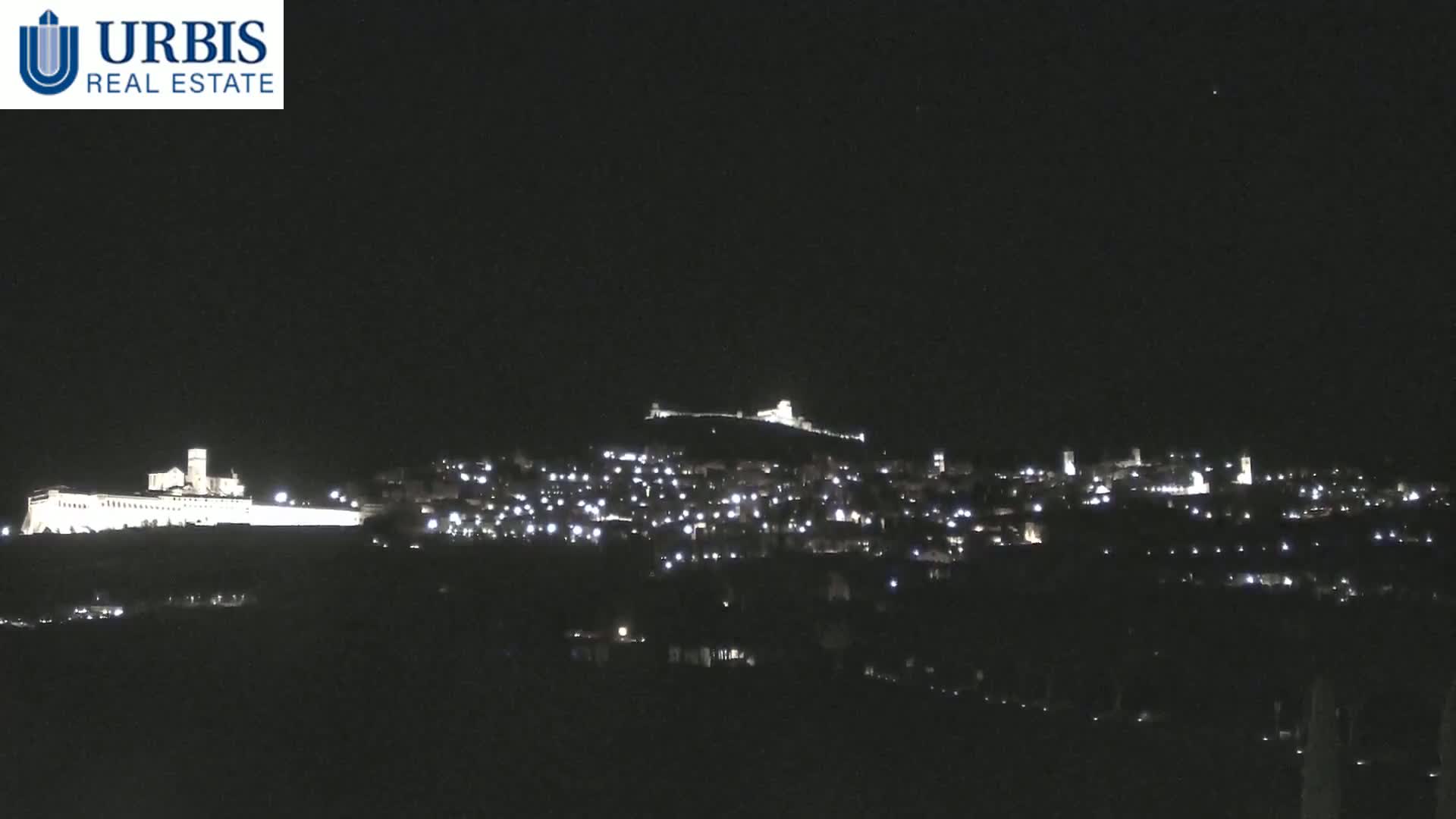 A nighttime long shot shows a hilltop town illuminated by many small lights, with a larger, brighter structure on the left and another on a hill in the center.