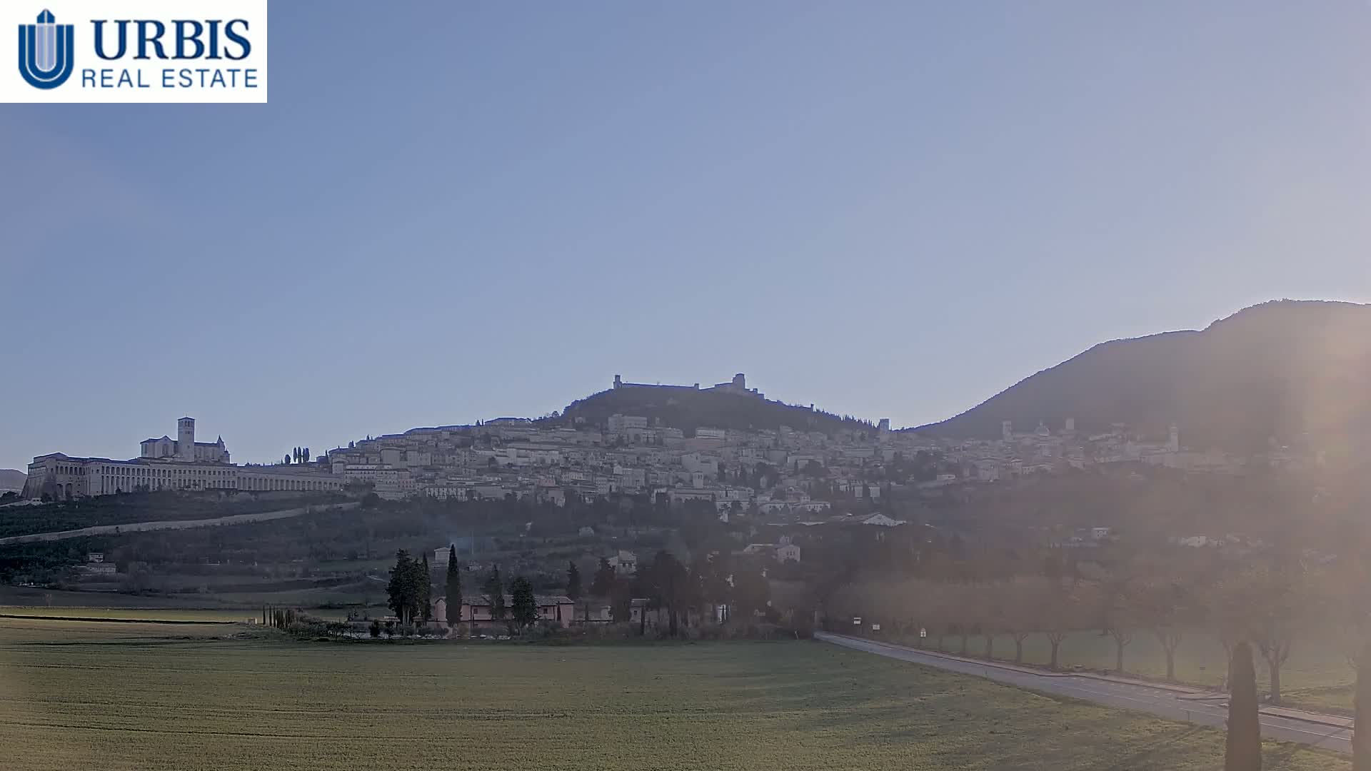 A hazy view of a hilltop town, with a large building complex prominently featured, stretches across a landscape under a clear, bright sky.