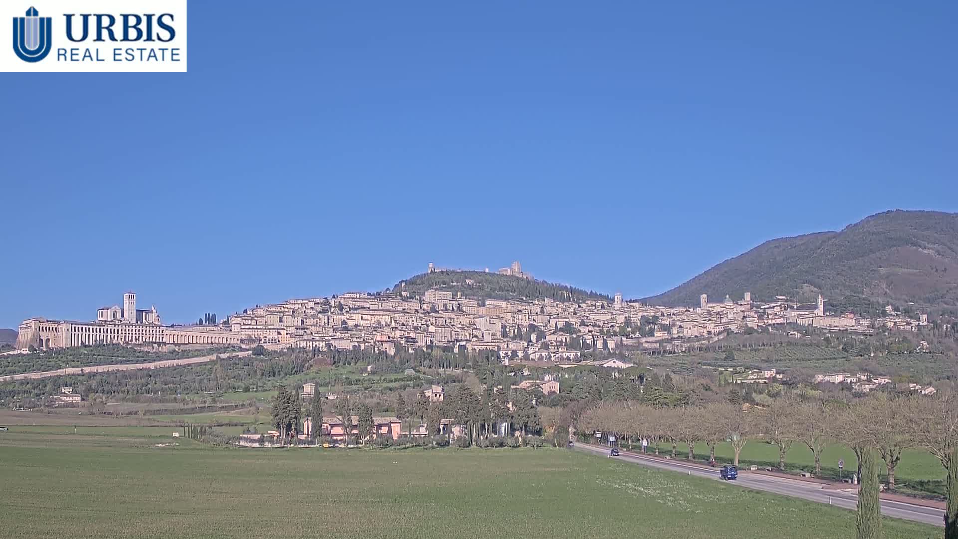 A sprawling hilltop town, dominated by large buildings, sits under a clear blue sky, with a flat, grassy plain in the foreground.
