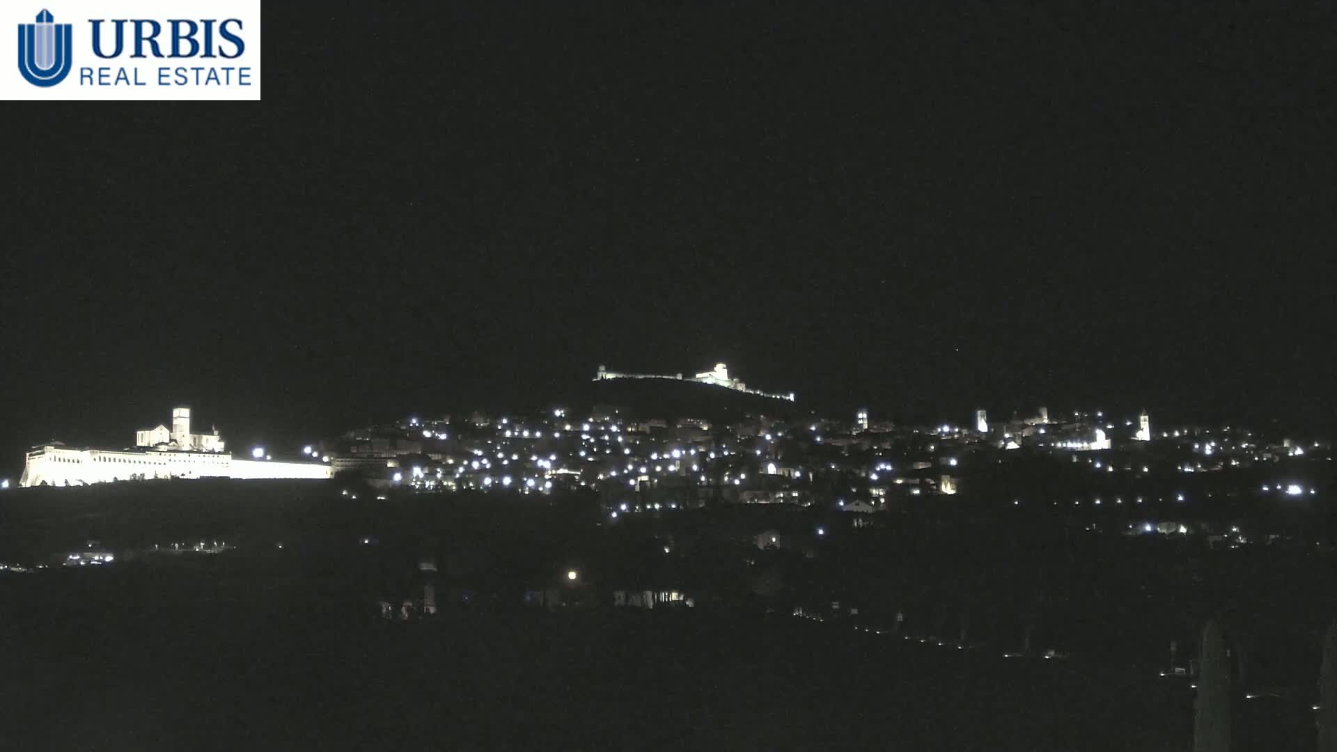 A nighttime view of a hilltop town illuminated by scattered lights, featuring a large, brightly lit building at the base of the hill.
