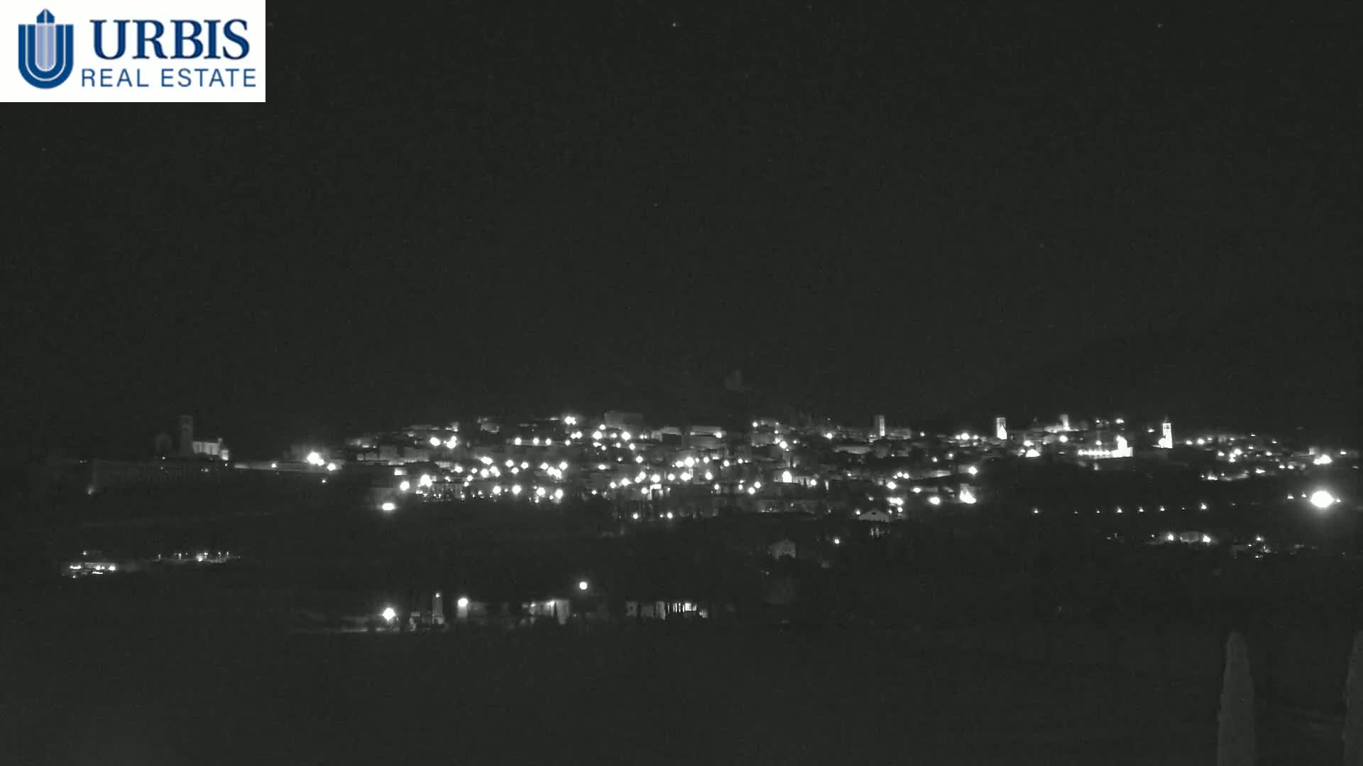 A nighttime long shot shows a small town illuminated by scattered lights on a hill under a clear, dark sky.