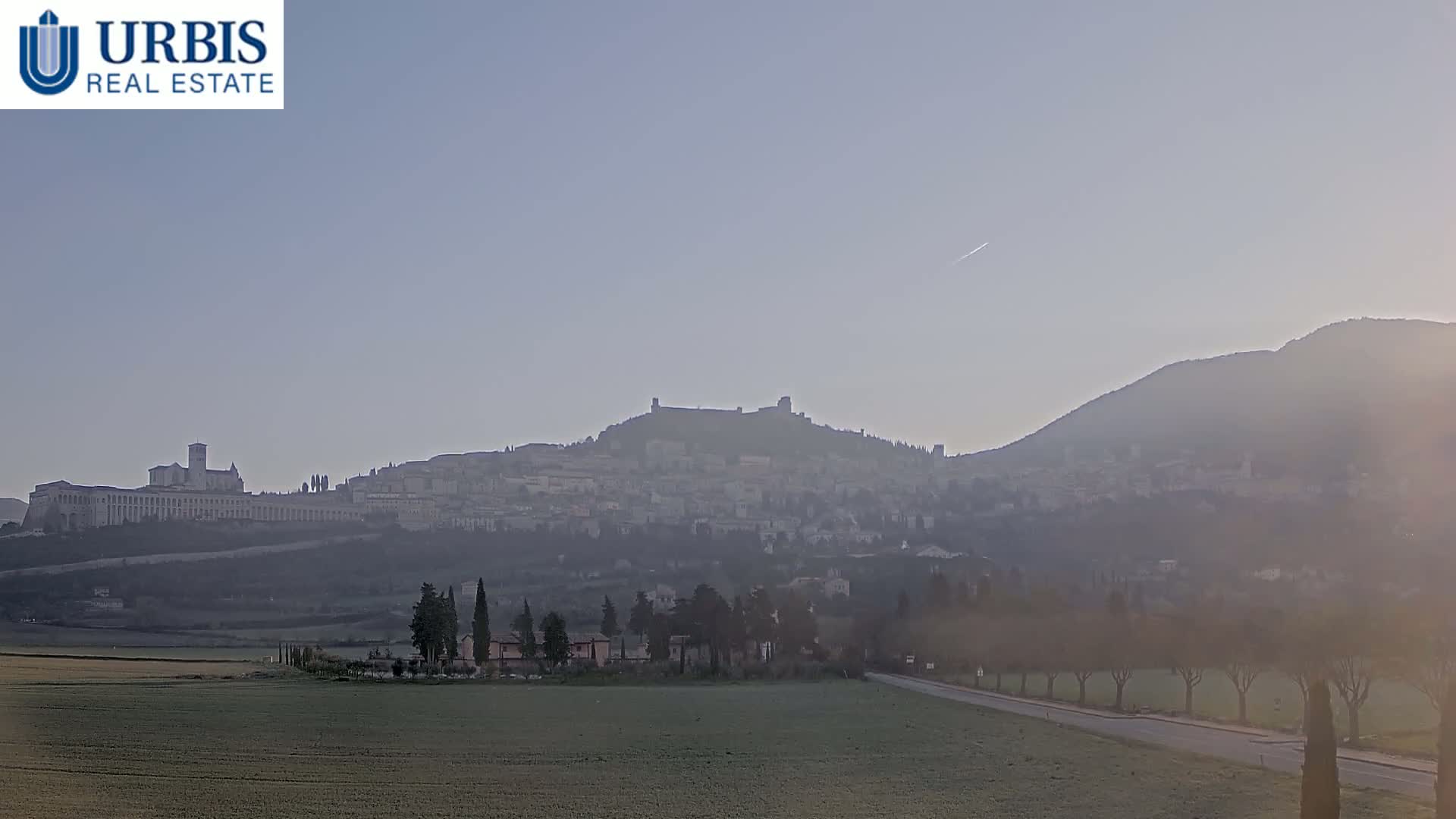 A hazy view shows a distant town nestled on a hill, with a larger structure possibly a fortress or castle, on a hilltop under a clear, light-blue sky.