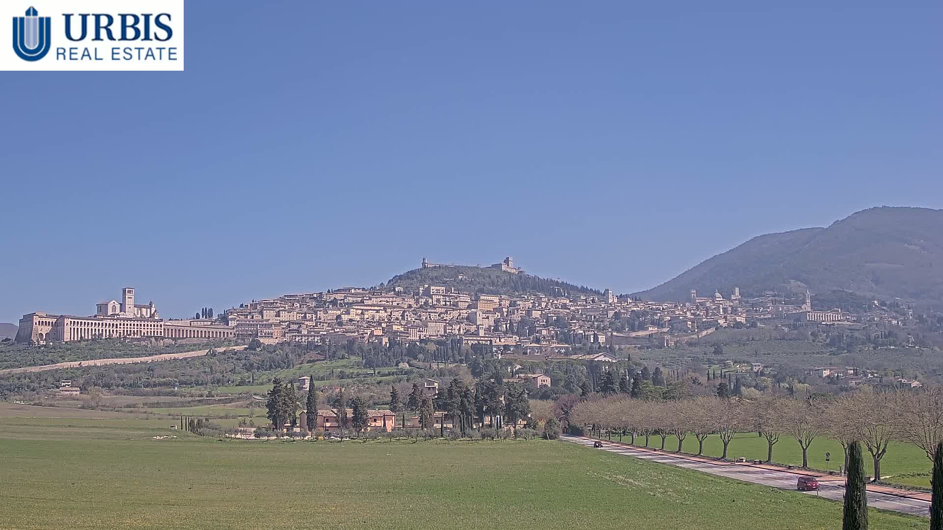 A large hilltop town is visible in the distance under a clear blue sky, with a long road and green fields in the foreground.