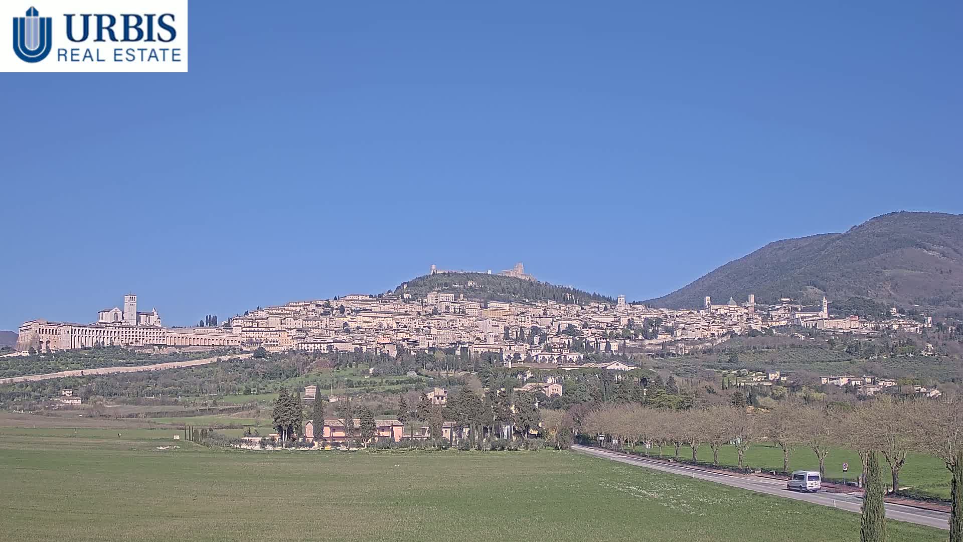 A large hill town, possibly Assisi, is visible under a clear blue sky, with a van driving on a road in the foreground.