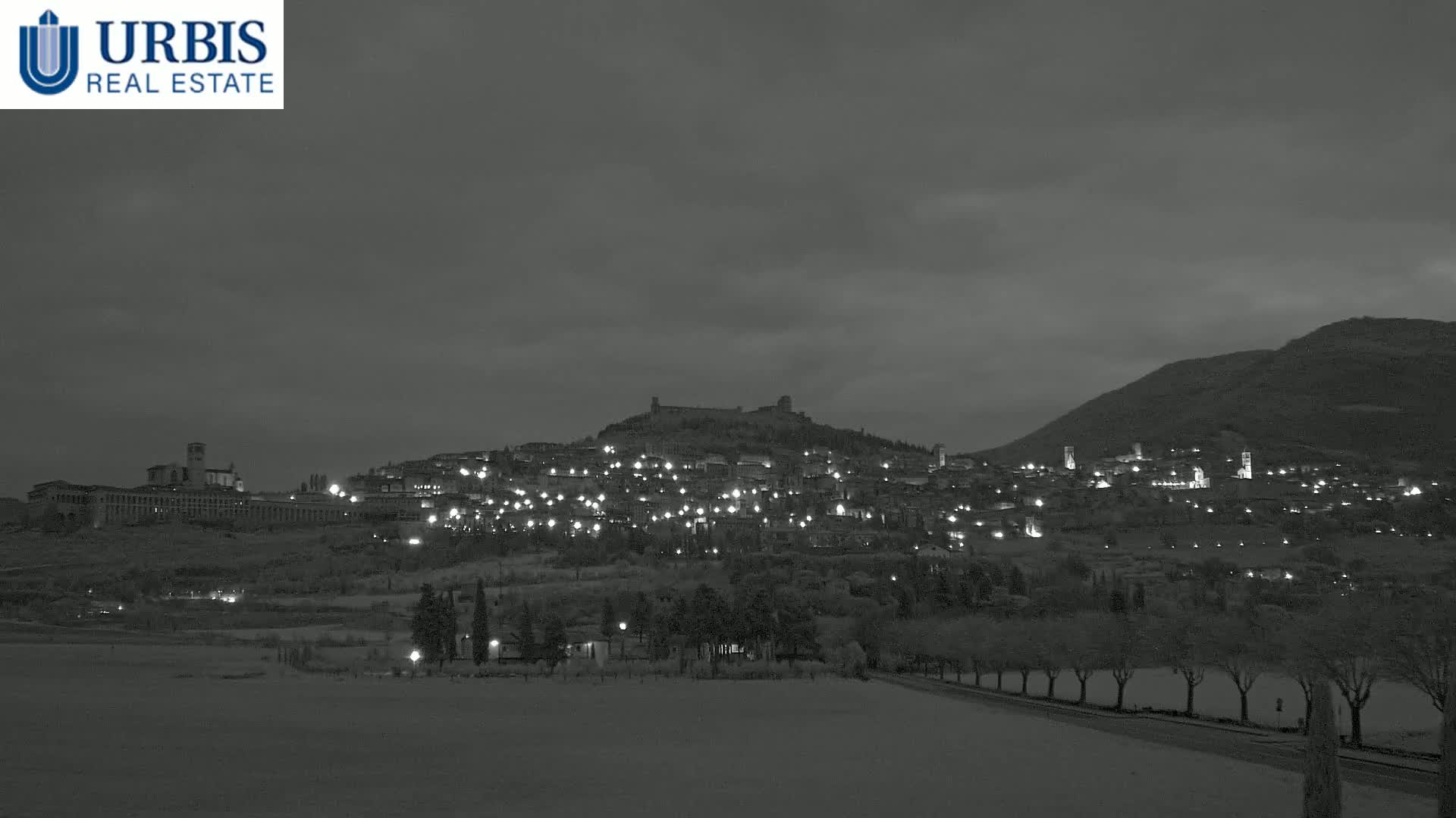 A hilltop town, illuminated at night, is nestled between two hills under a cloudy, dark sky.