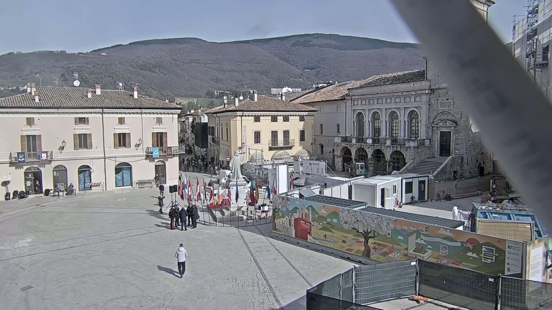 A town square, under a clear sky, features several buildings, a monument, and temporary structures, with a few people present.