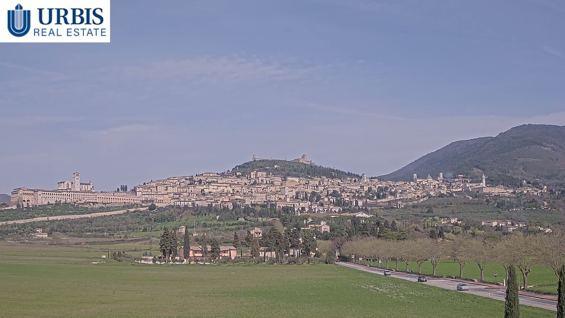 A sprawling hilltop town, with a large basilica visible, is seen from a distance on a clear, sunny day, with a green field and road in the foreground.