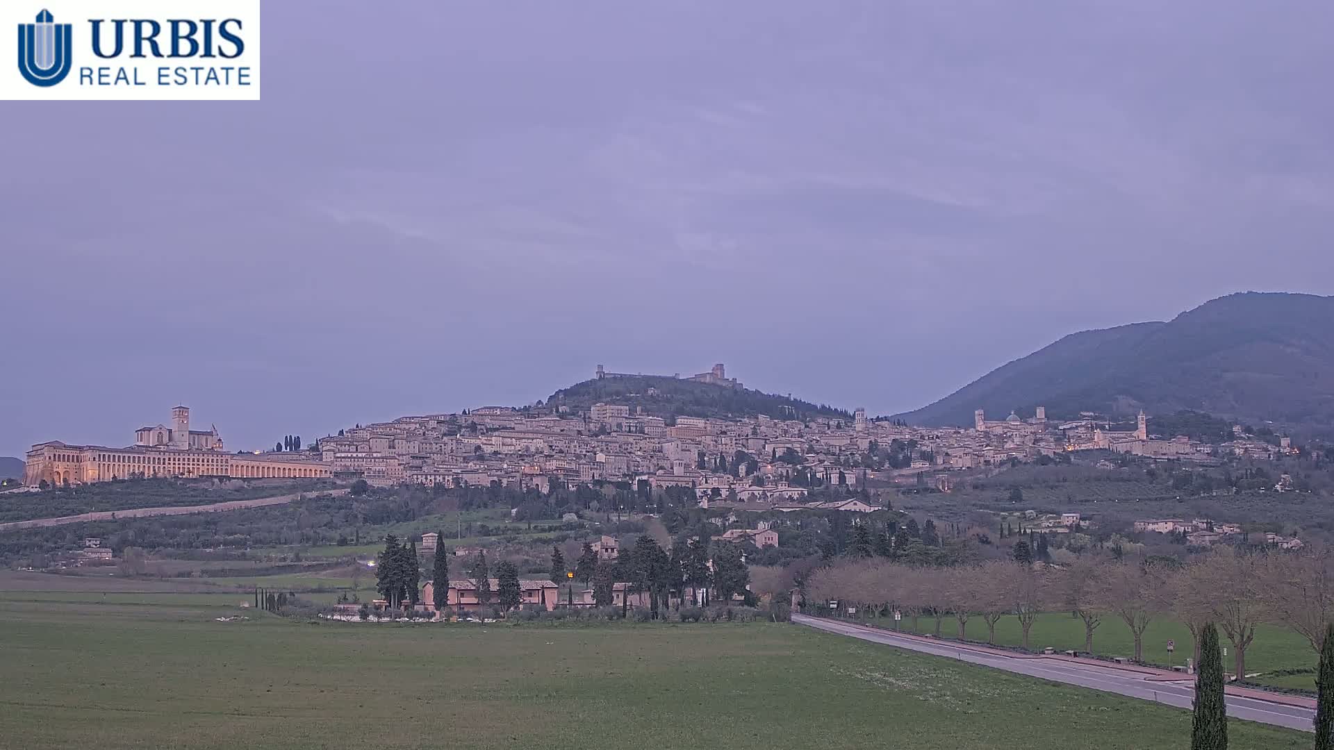 A hilltop town with numerous buildings and a large basilica is visible in the distance under a pale purple twilight sky, with a flat green field and road in the foreground.