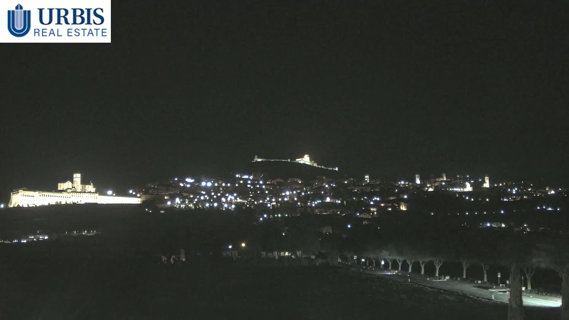 A nighttime view of a hilltop town, illuminated by lights, with a large building complex at the base of the hill.