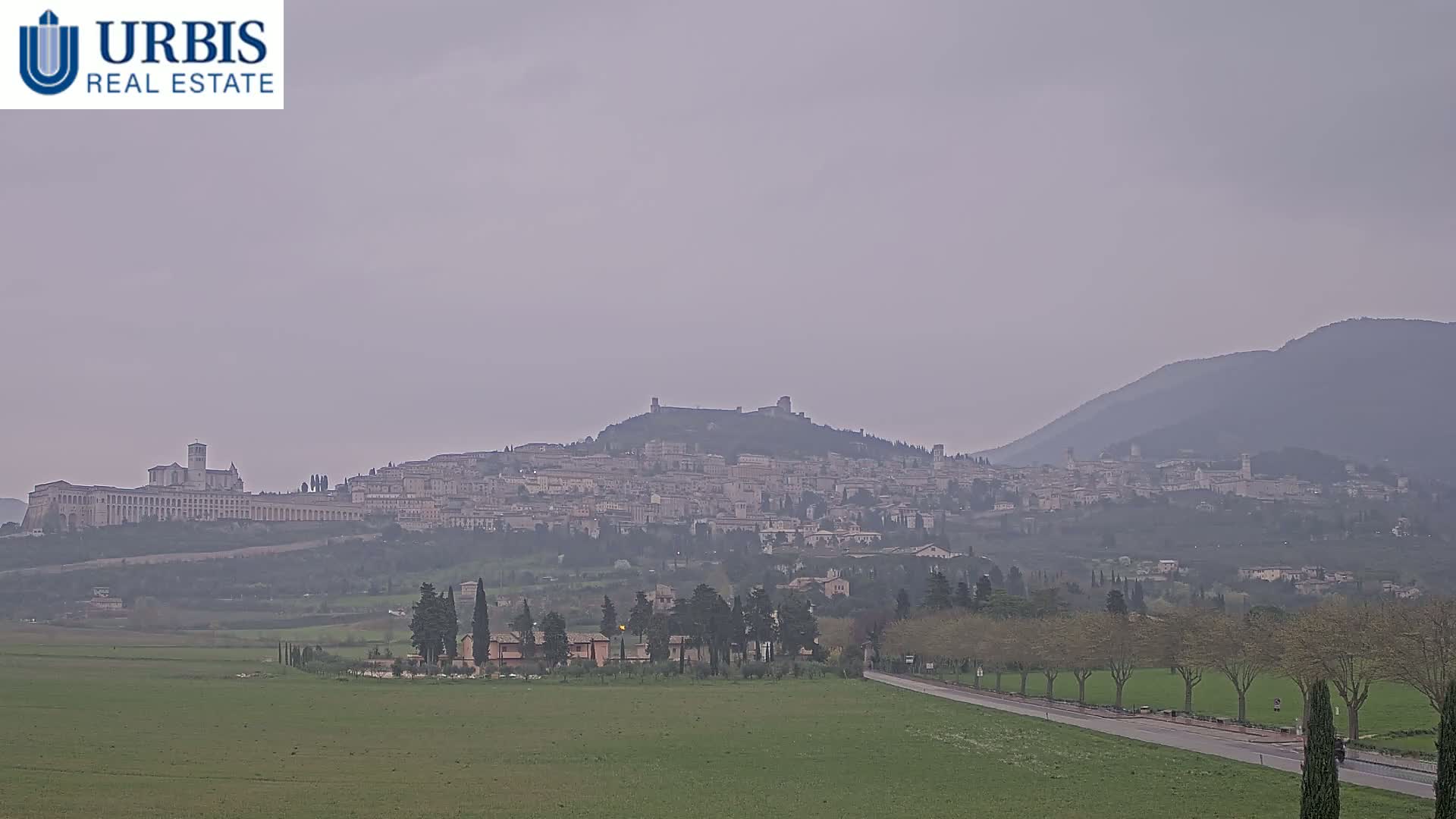 A hazy view shows a hilltop town nestled in a valley, with mountains in the background and a flat green field in the foreground.