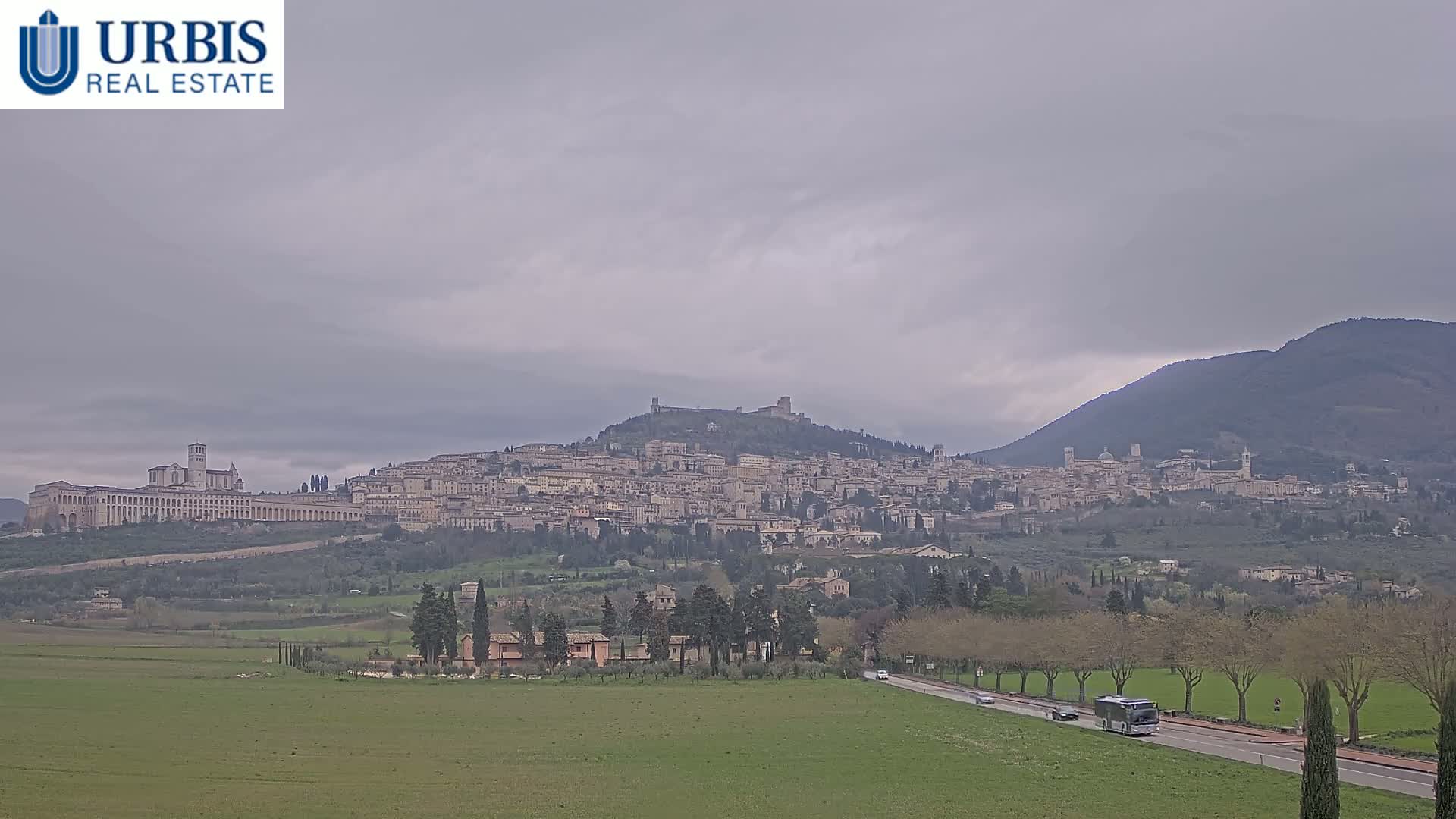 A large hill town with numerous buildings, including a large basilica, sits under a cloudy sky, with a road and green fields in the foreground.