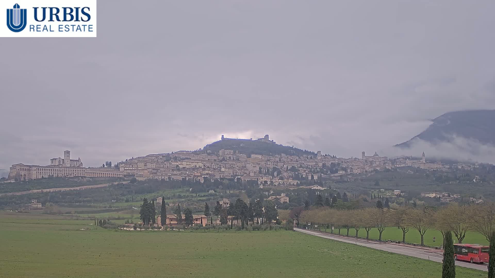 A hilltop town with many buildings, including a large basilica, is visible in the distance under an overcast sky; a red bus travels on a road in the foreground.