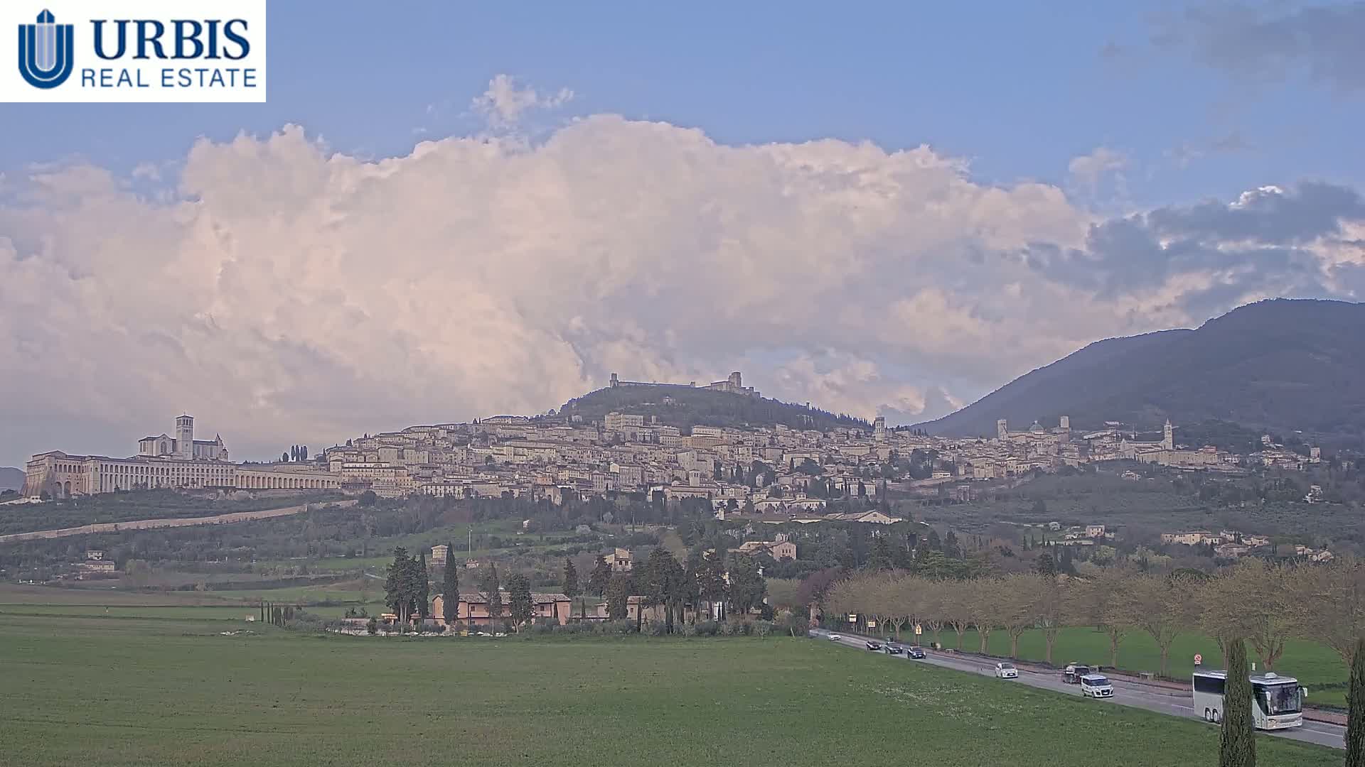A hilltop town with numerous buildings, including a large church or basilica, is visible under a partly cloudy sky, with a road and flat farmland in the foreground.