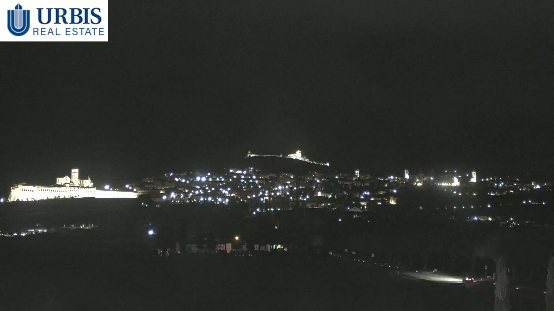 A nighttime long shot shows a hilltop town illuminated, with a large, brightly lit building at the base of the hill.