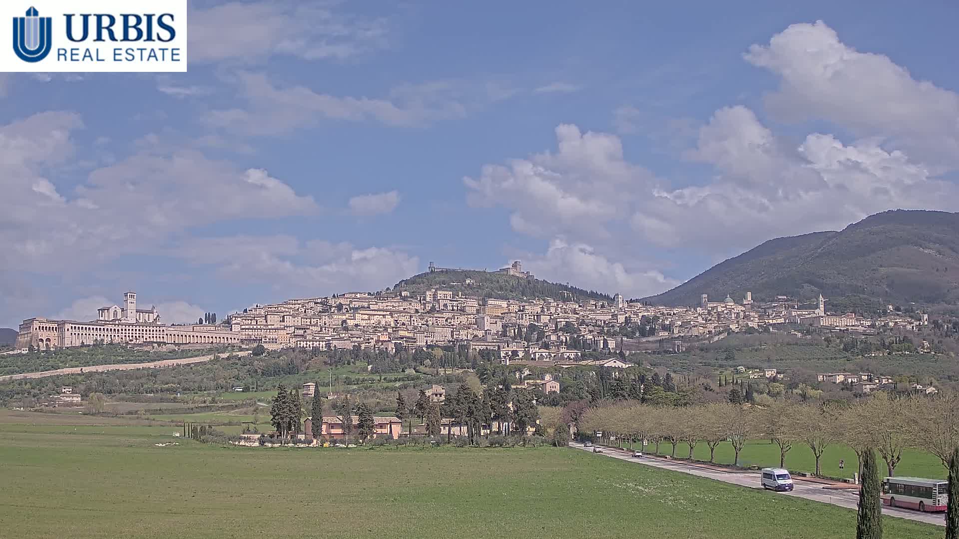 A large hilltop town is visible under a partly cloudy sky, with a van and bus driving on a road in the foreground.