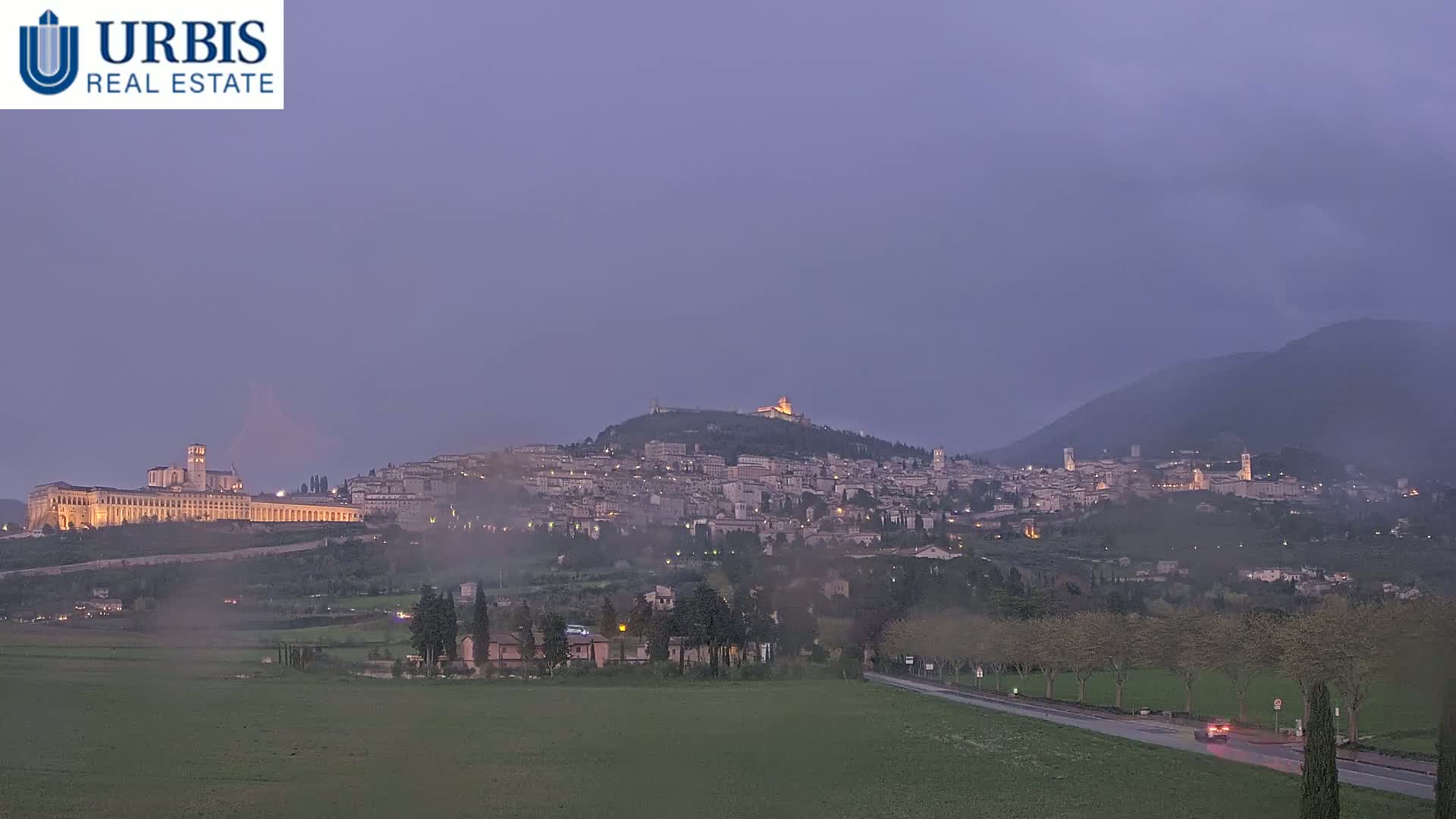 A hilltop town, illuminated at dusk, is nestled between a large flat field and a mountain range under a hazy purple sky.