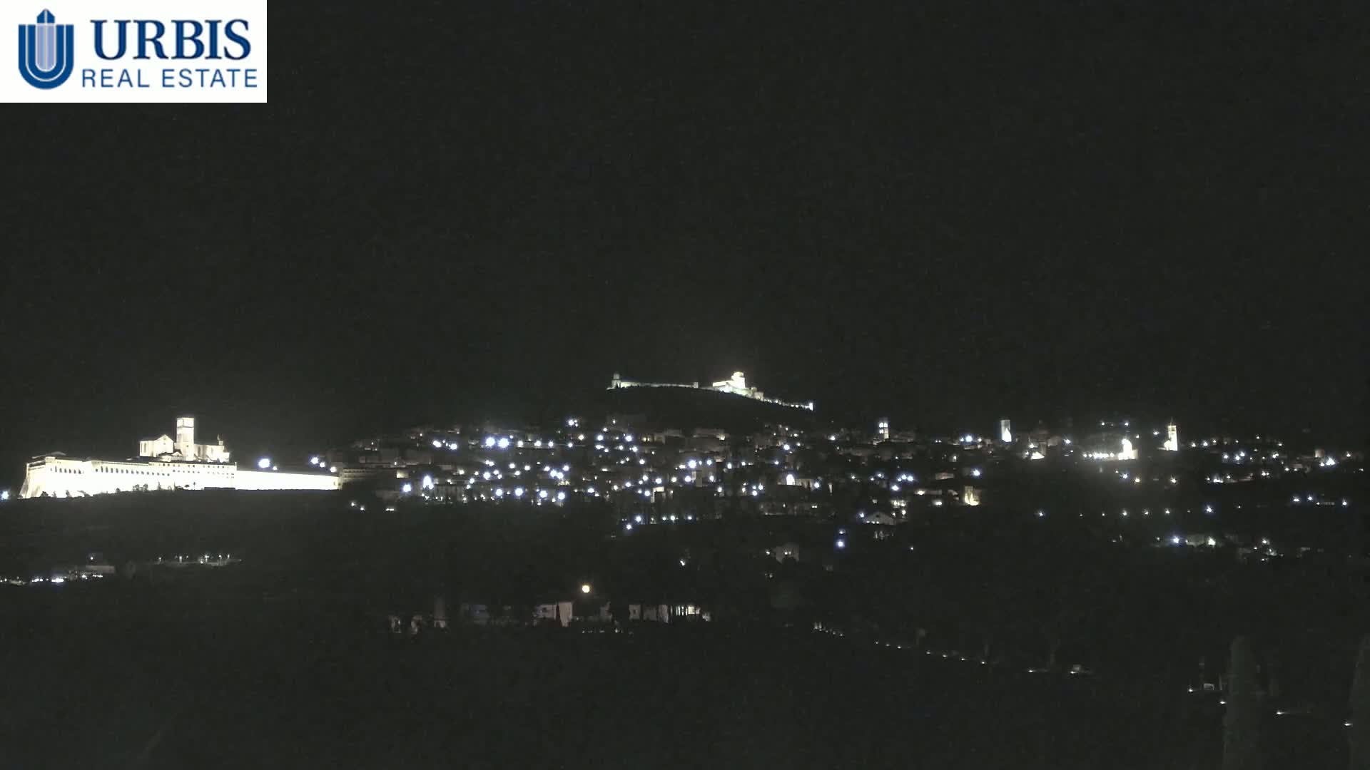 A nighttime view of a hilltop town illuminated by numerous lights, with a large, brightly lit structure visible at the base of the hill.