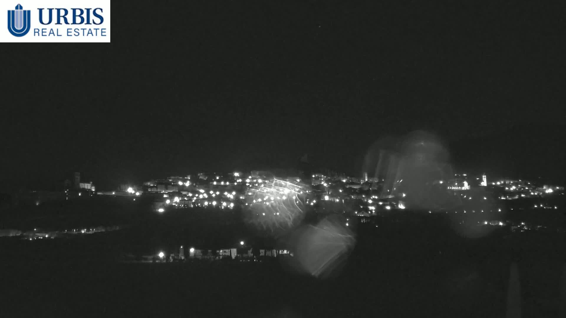 A nighttime long shot of a town illuminated by scattered lights, viewed through rain-streaked glass under a dark sky.