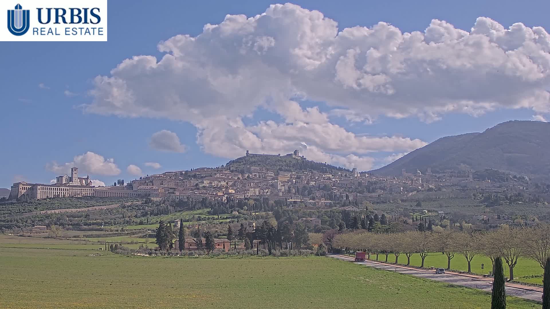 A hilltop town, with a large basilica visible, sits under a partly cloudy sky, overlooking a flat green field with a road and trees.