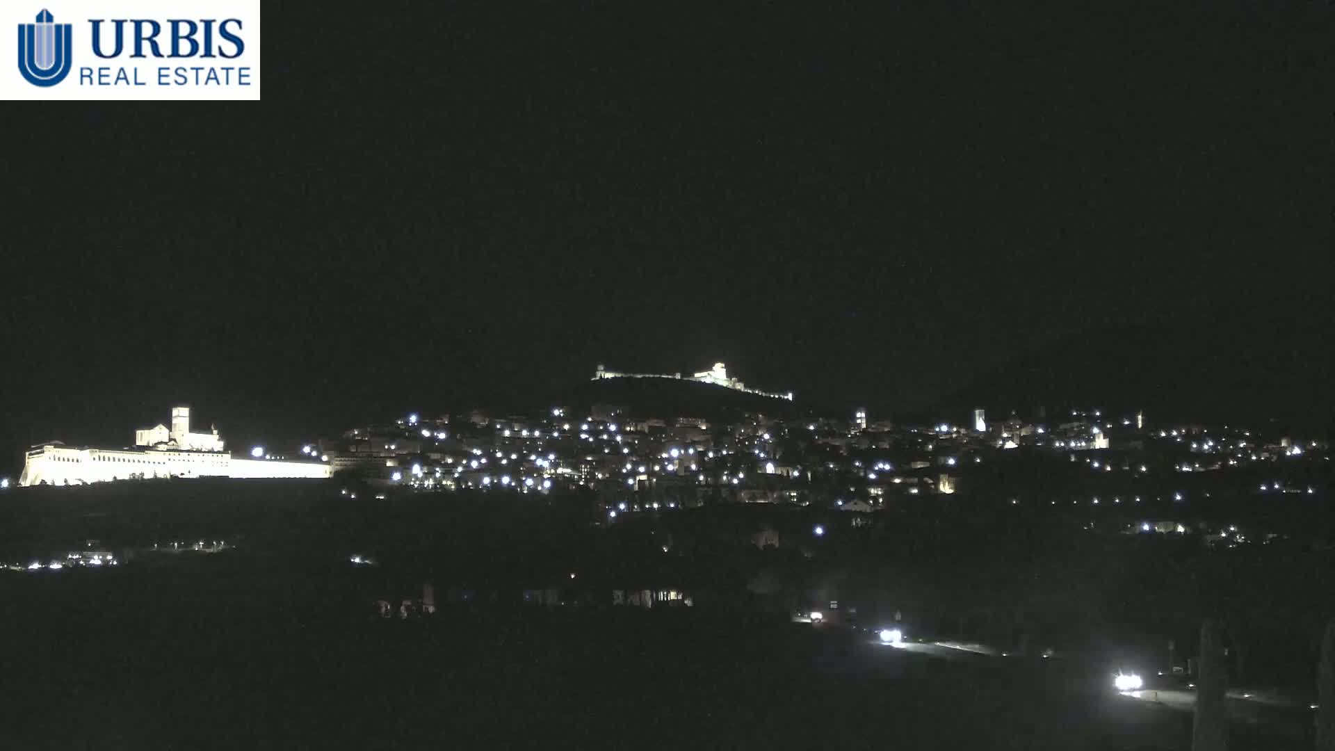 A nighttime view of a hilltop town illuminated by lights, with a large, brightly lit building on a lower hill in the foreground under a clear, dark sky.
