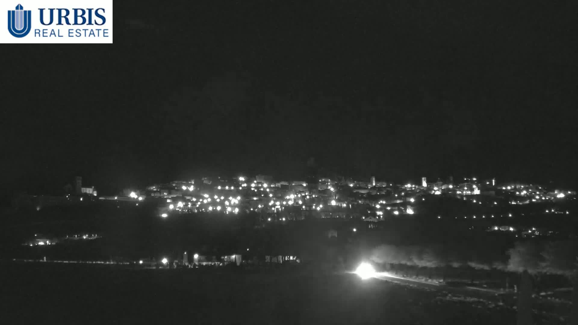 A nighttime long shot shows a town illuminated by its lights, with a bright light source in the foreground on a dark road.