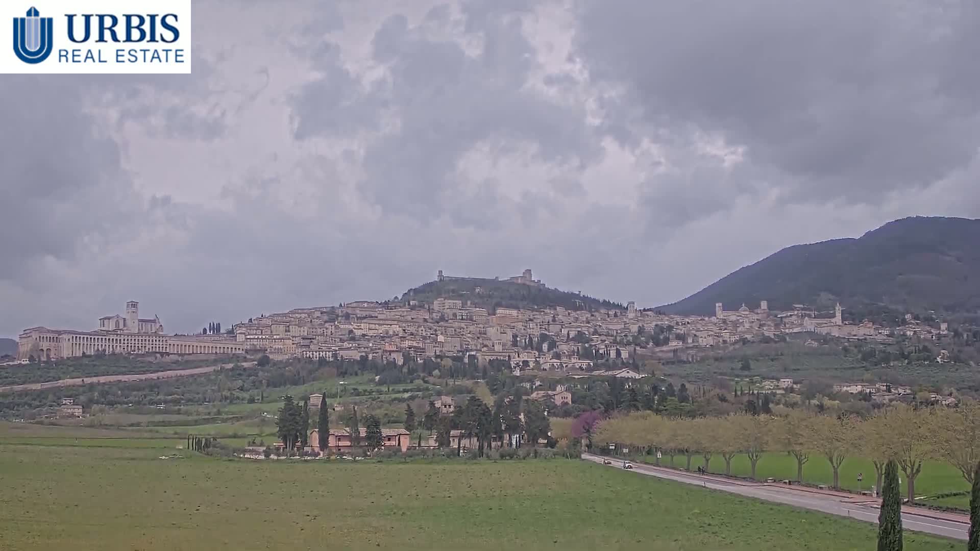 A hilltop town, partially shrouded in overcast skies, sits nestled between rolling green hills and a larger mountain range in the distance.