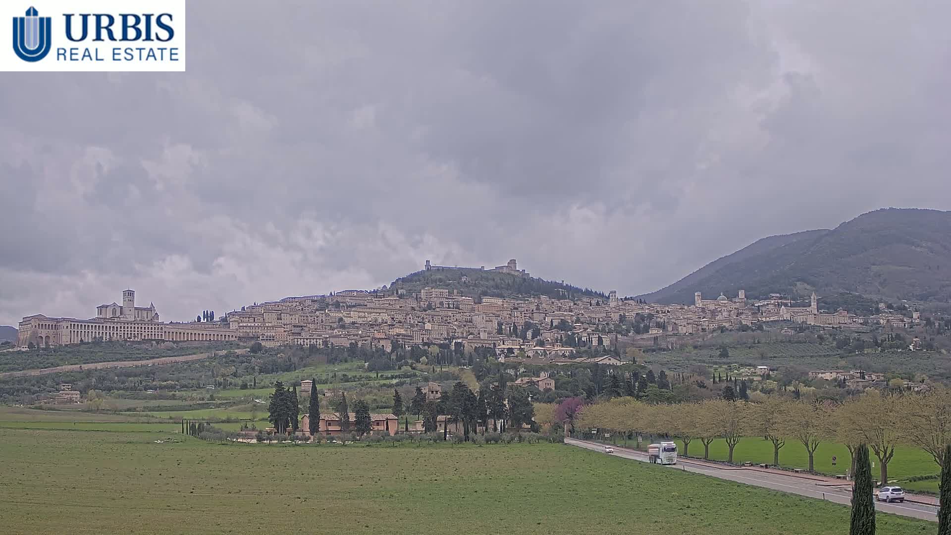 A large hilltop town, under a cloudy sky, is visible from a distance across a flat, grassy plain with a road and sparse trees.