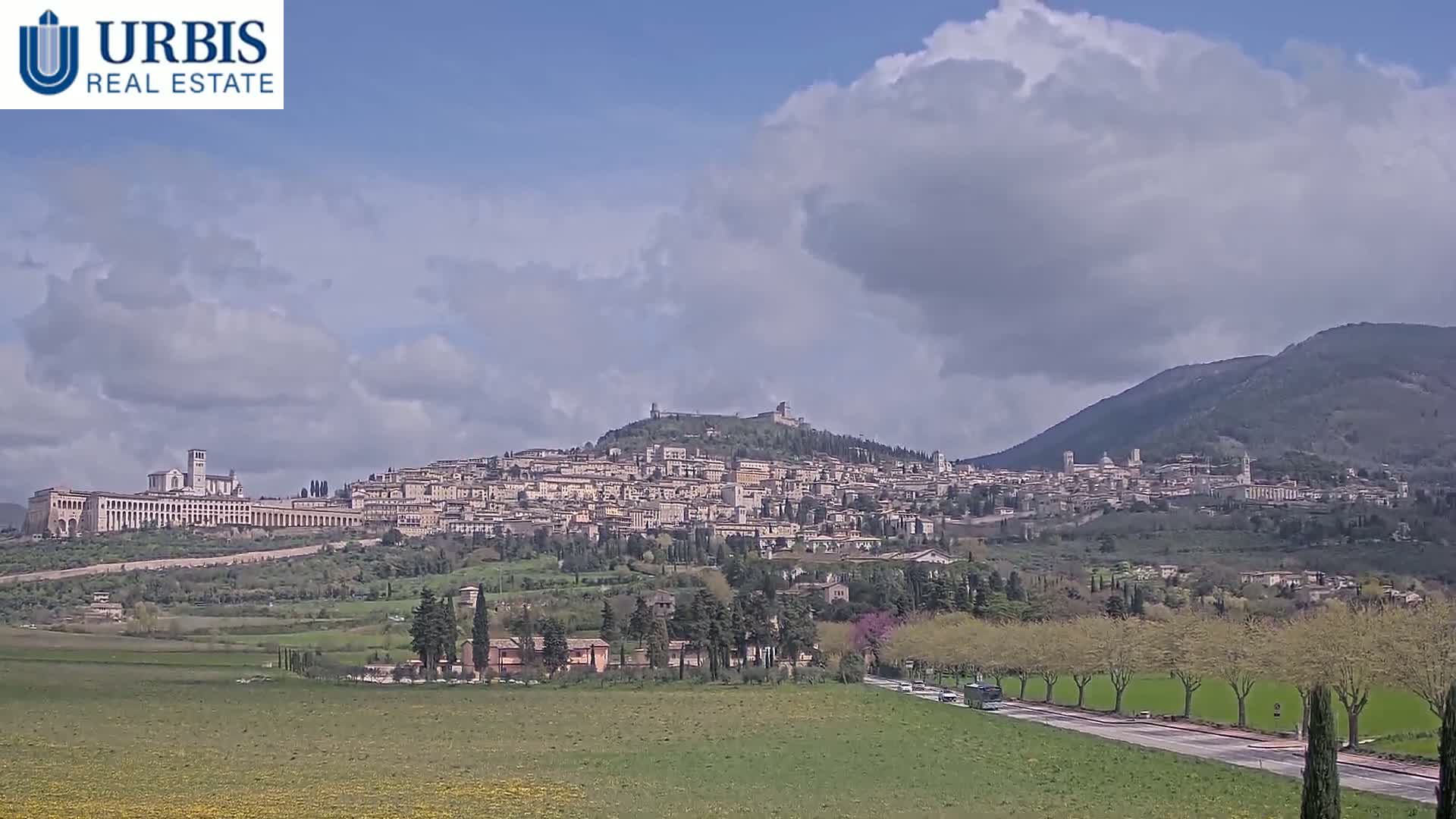 A hilltop town with a large basilica, nestled in a valley under a partly cloudy sky.