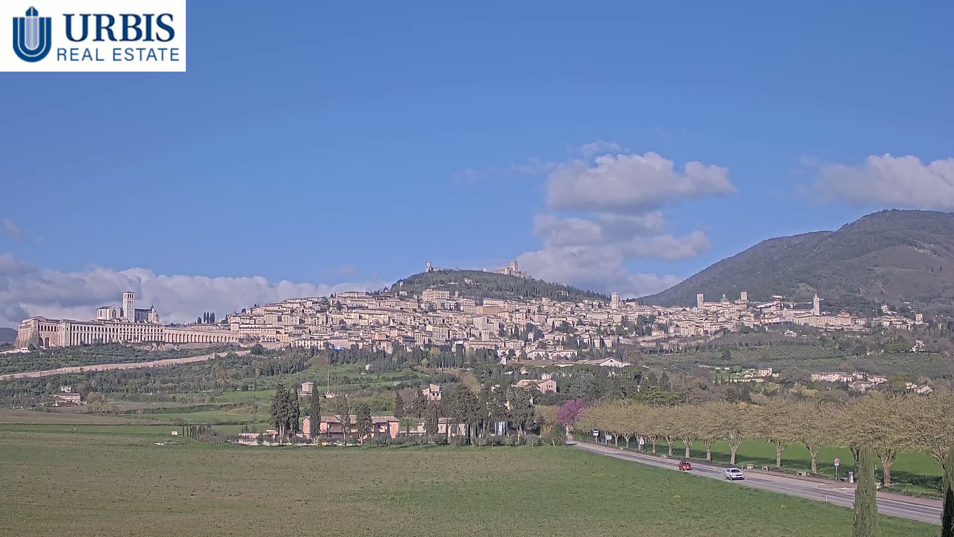 A hilltop town, under a partly cloudy blue sky, is viewed from a distance across a flat field with a road and trees in the foreground.