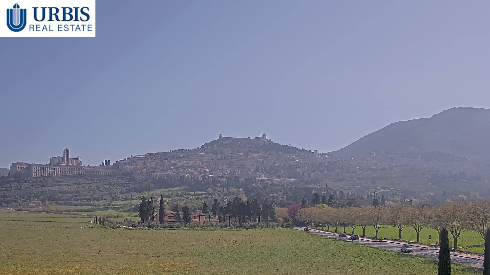 A hilltop town, partially obscured by haze, is visible in the distance, with a long, flat road lined with trees leading towards it across a green field under a clear sky.