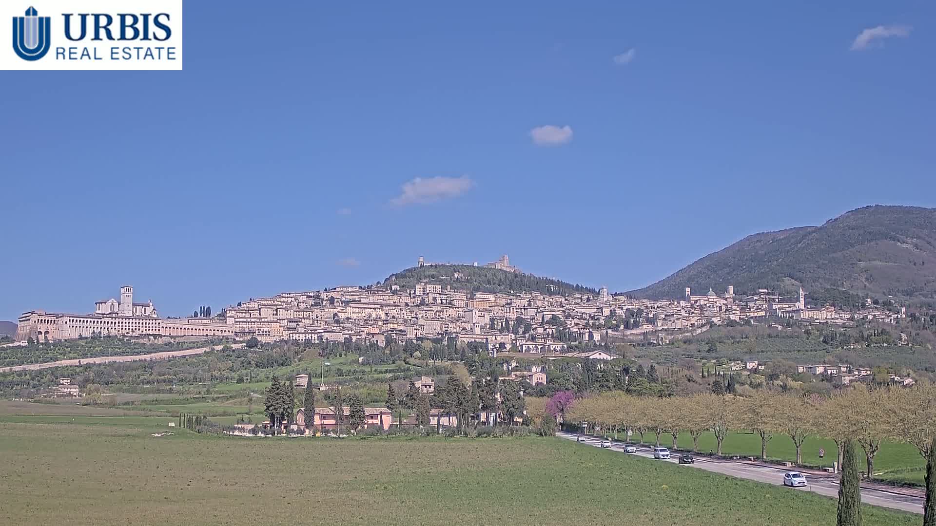 A hilltop town, filled with buildings of similar style and color, sits under a clear blue sky with a few scattered clouds, and a road with sparse traffic leads toward the town from a grassy field in the foreground.