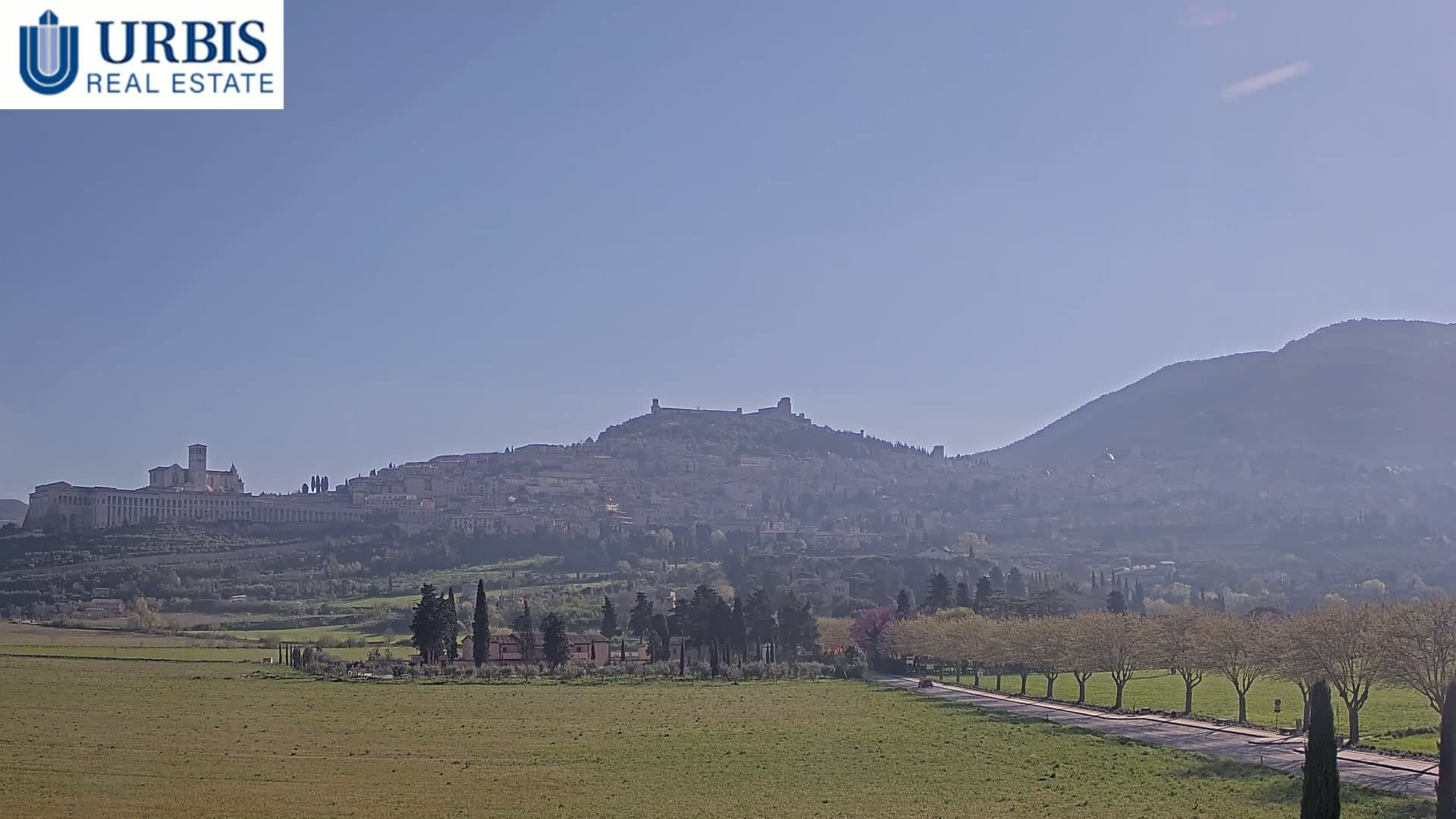 A hilltop town, partially obscured by haze, sits below a larger mountain under a clear, bright sky.