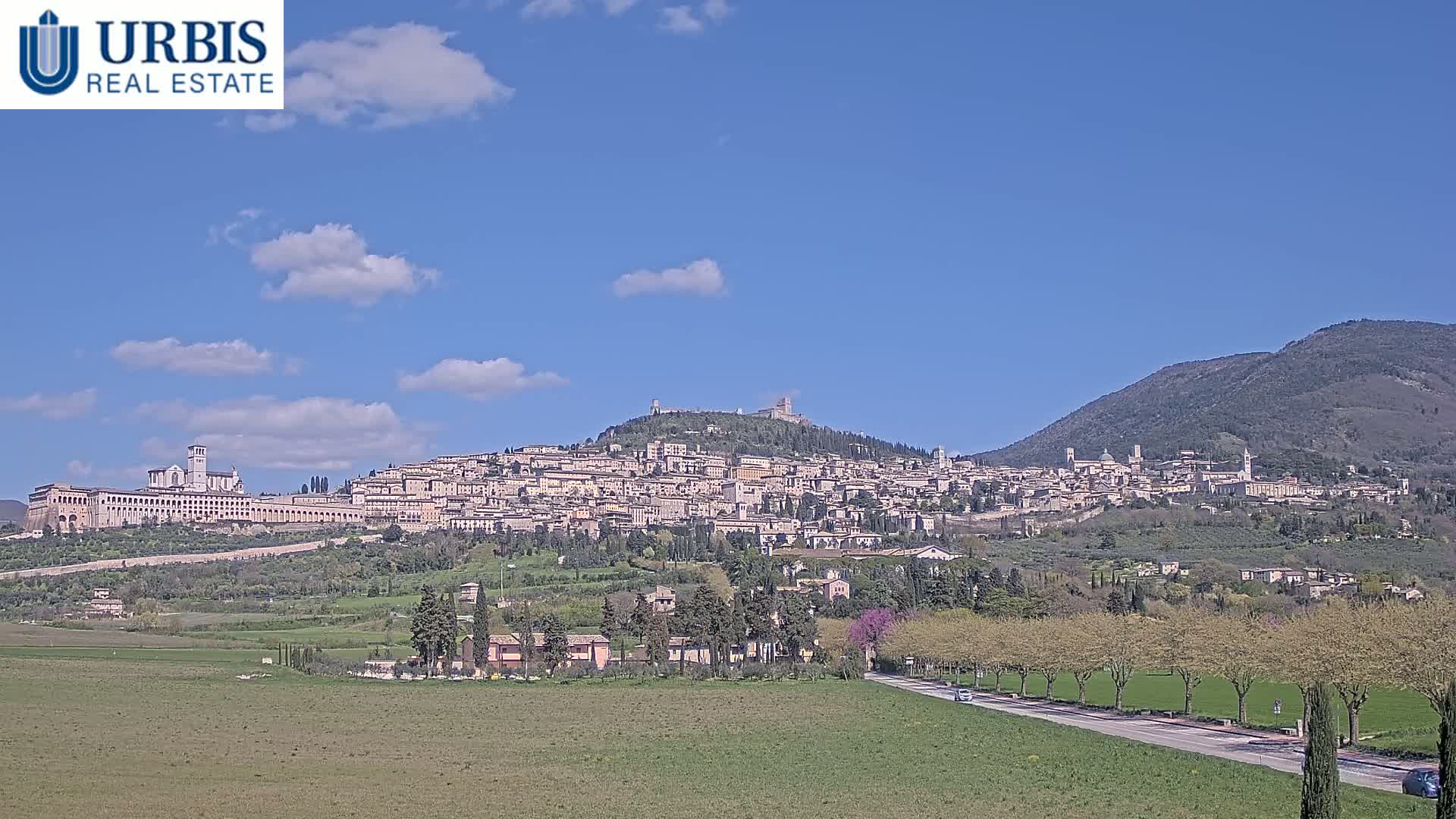 A hilltop town with numerous buildings, including a large church, is visible under a mostly clear blue sky with a few clouds, situated next to a large mountain and a flat green field in the foreground.