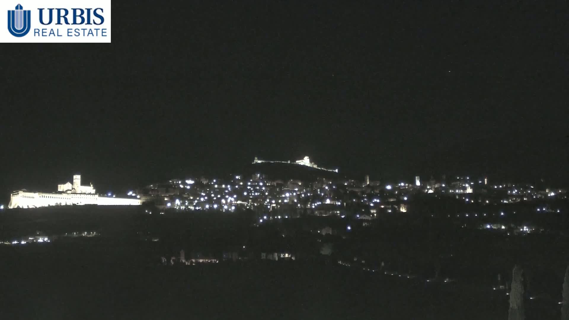 A nighttime long shot shows a hilltop town with lights illuminating buildings, including a large structure that looks like a basilica, under a clear, dark sky.