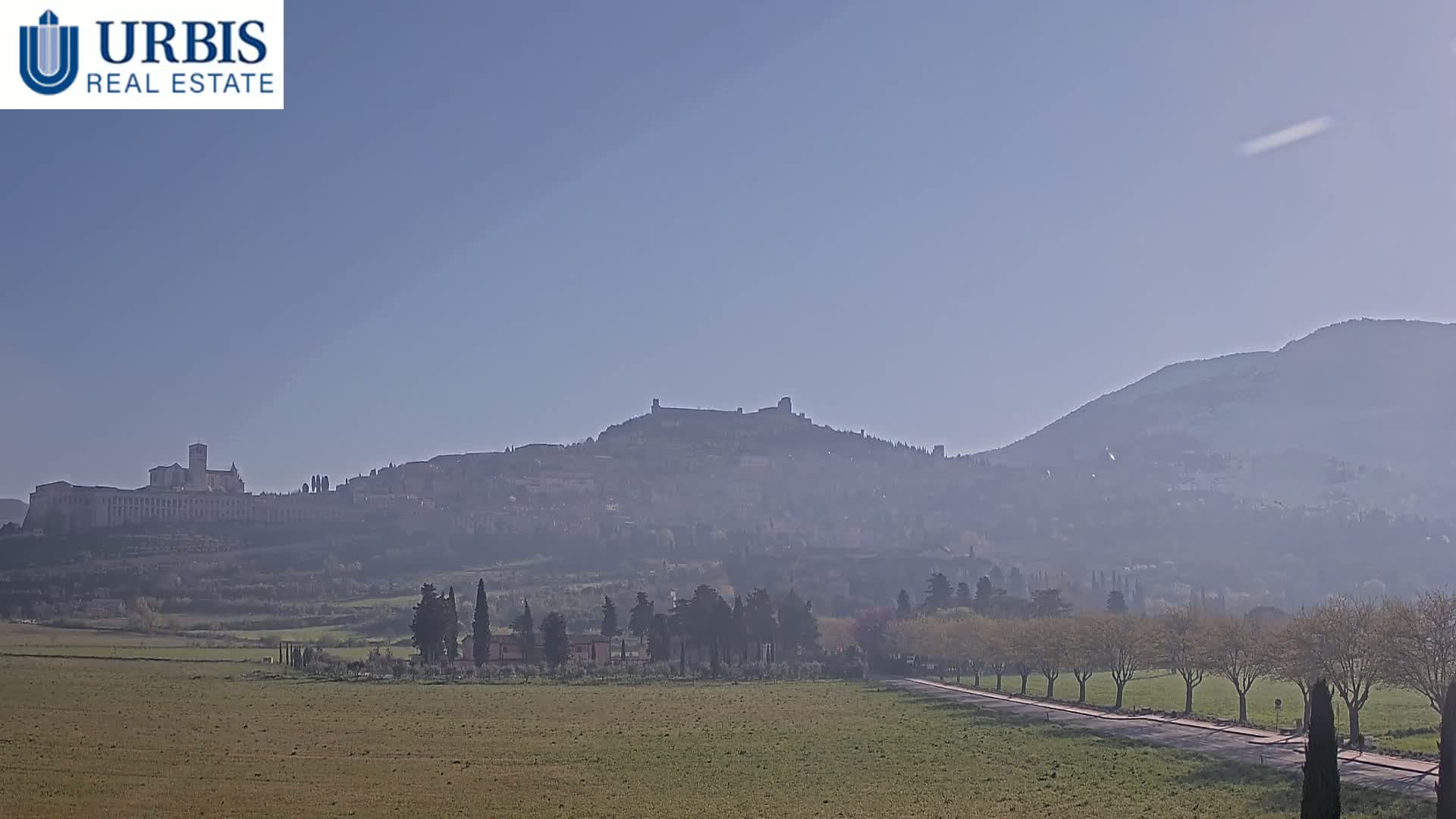 A hazy, distant view of a hilltop town, including a large stone structure, under a clear, pale blue sky.