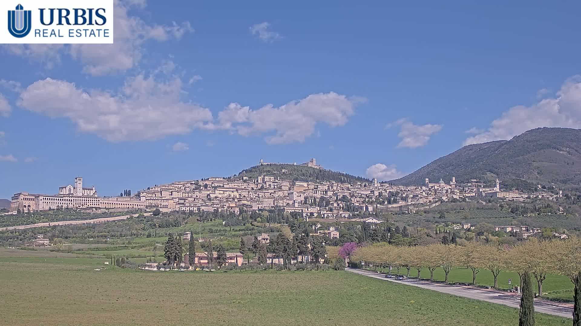 A hilltop town with many buildings, including a large basilica, is visible under a mostly clear blue sky with some scattered clouds, and sits nestled in a green valley between two mountains.