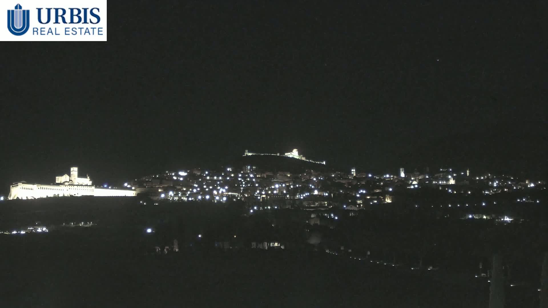 A nighttime long shot shows a hilltop town illuminated by numerous lights, with a large, brightly lit building on a lower hill in the foreground.
