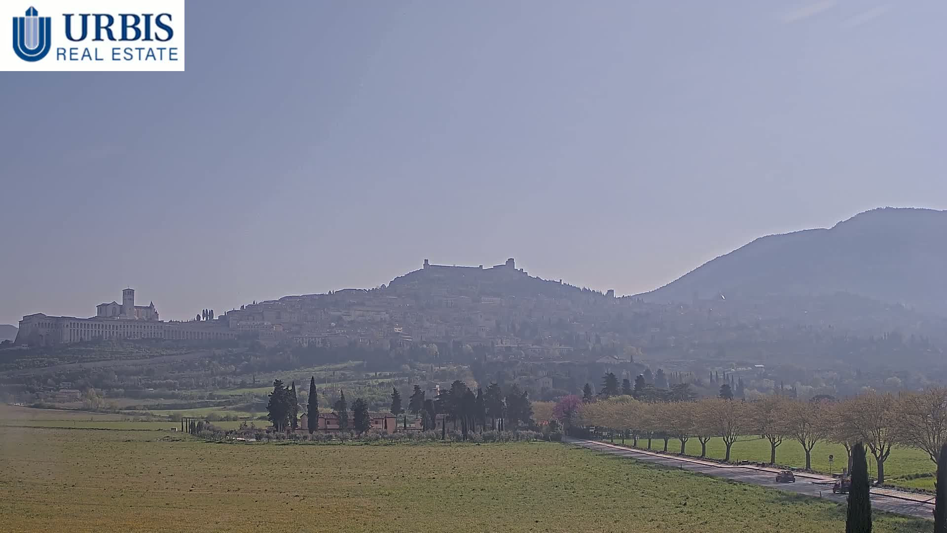 A hazy view shows a hilltop town with a fortress, surrounded by fields and trees under a clear sky.