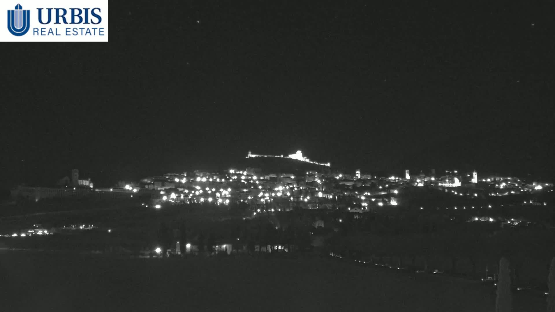 A nighttime long shot shows a hilltop town illuminated by many small lights under a clear, dark sky.