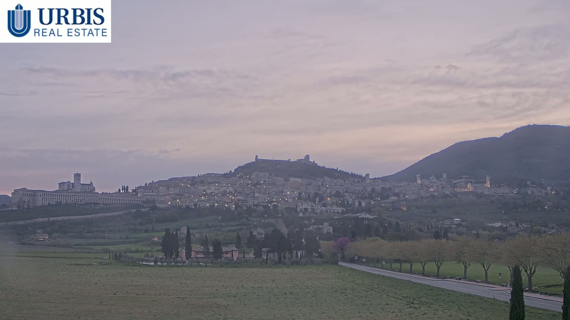 A hilltop town, illuminated at dusk under a pale, cloudy sky, sits nestled between a large, flat field and a distant mountain.