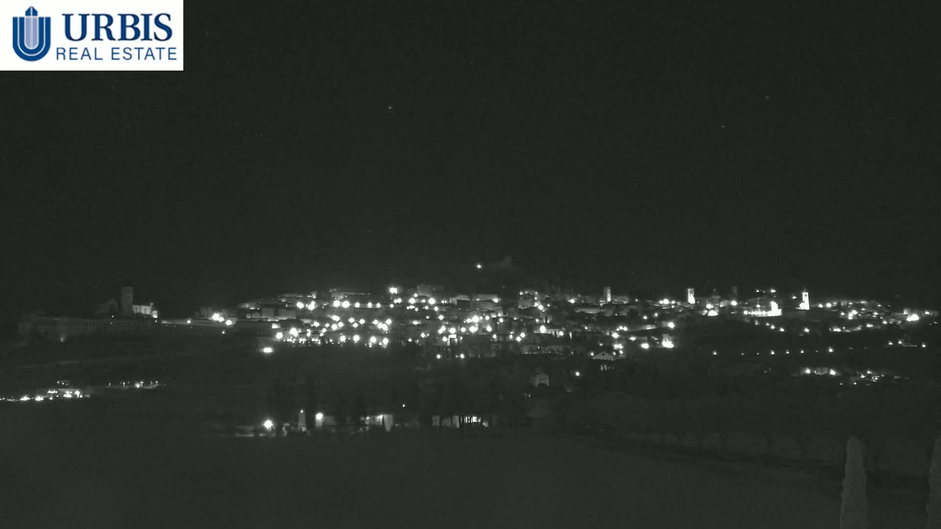 A nighttime, long shot of a small town illuminated by lights on a hill, under a clear, dark sky.