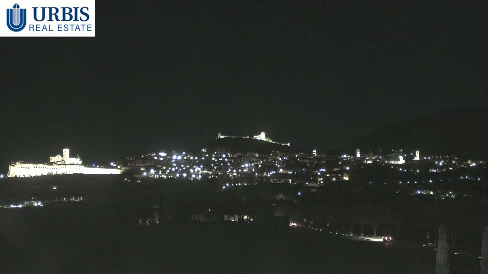 A nighttime view of a town nestled on a hill, illuminated by scattered lights, with a larger, brightly lit structure on a hill in the background.