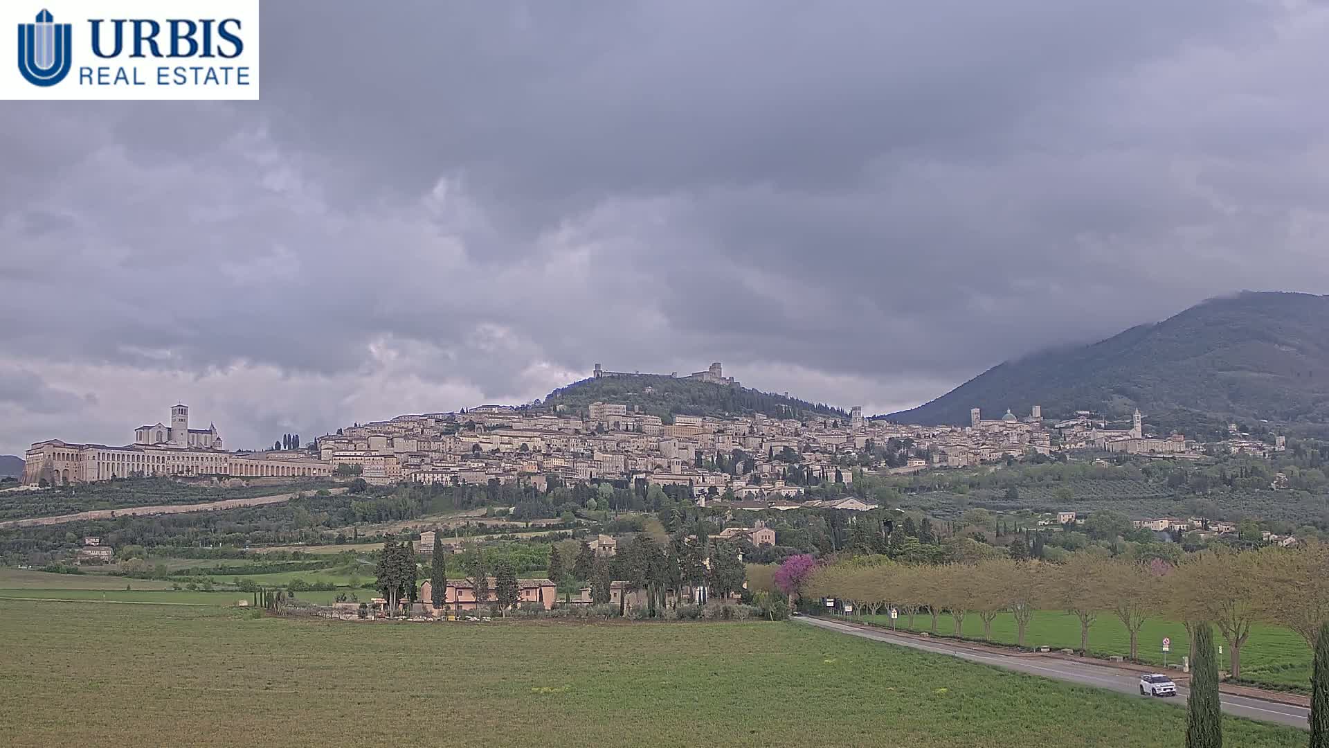 A large hill town, under a cloudy sky, sits nestled between two mountains and is viewed from a distance across a flat expanse of green fields.
