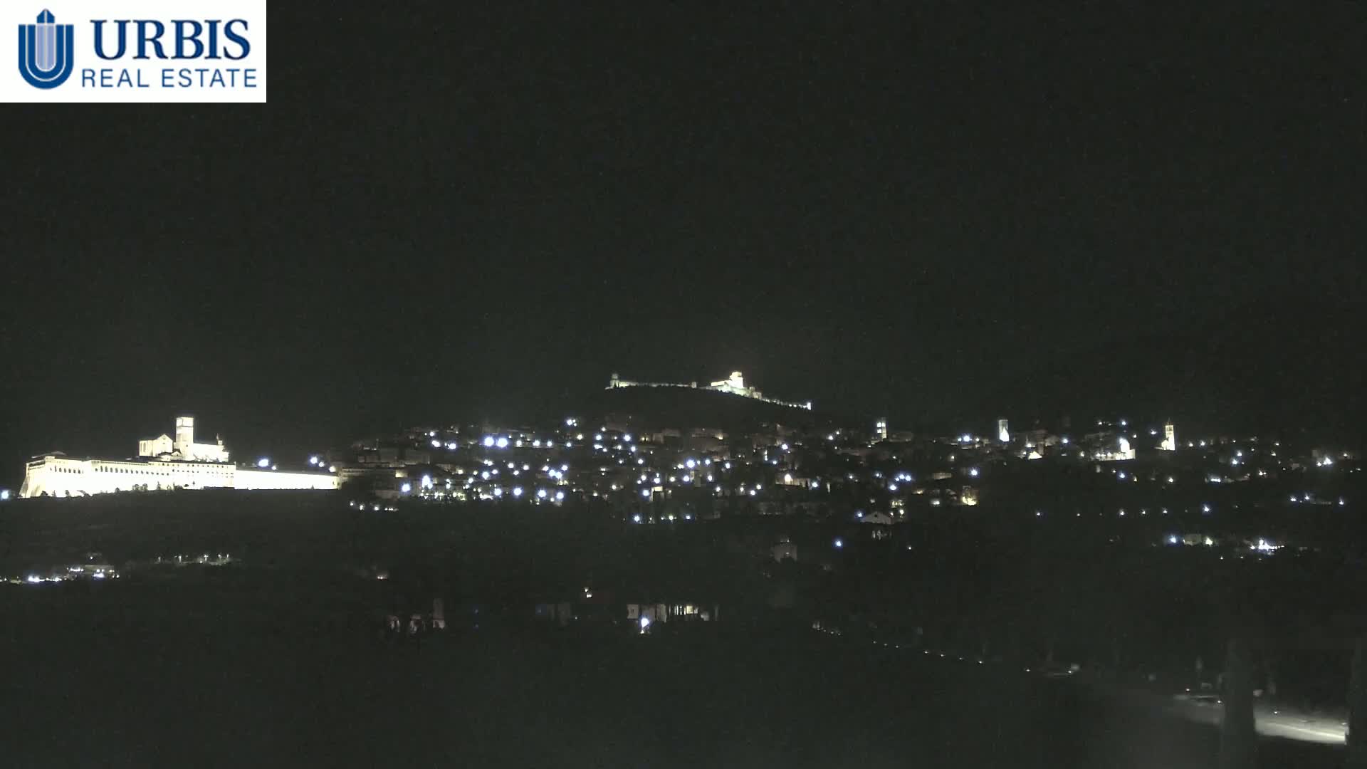 A hilltop town is illuminated at night, with a large, brightly lit building on a lower hill in the foreground.