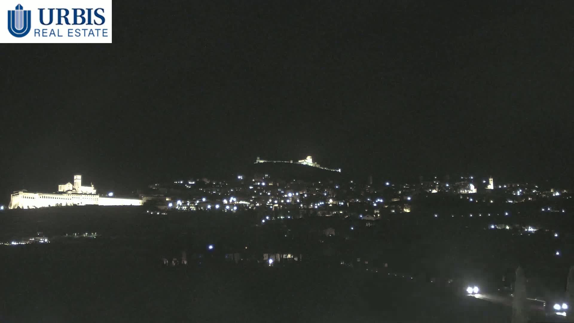 A nighttime view of a town nestled on a hill, illuminated by scattered lights, with a large, brightly lit structure on a hill in the background.