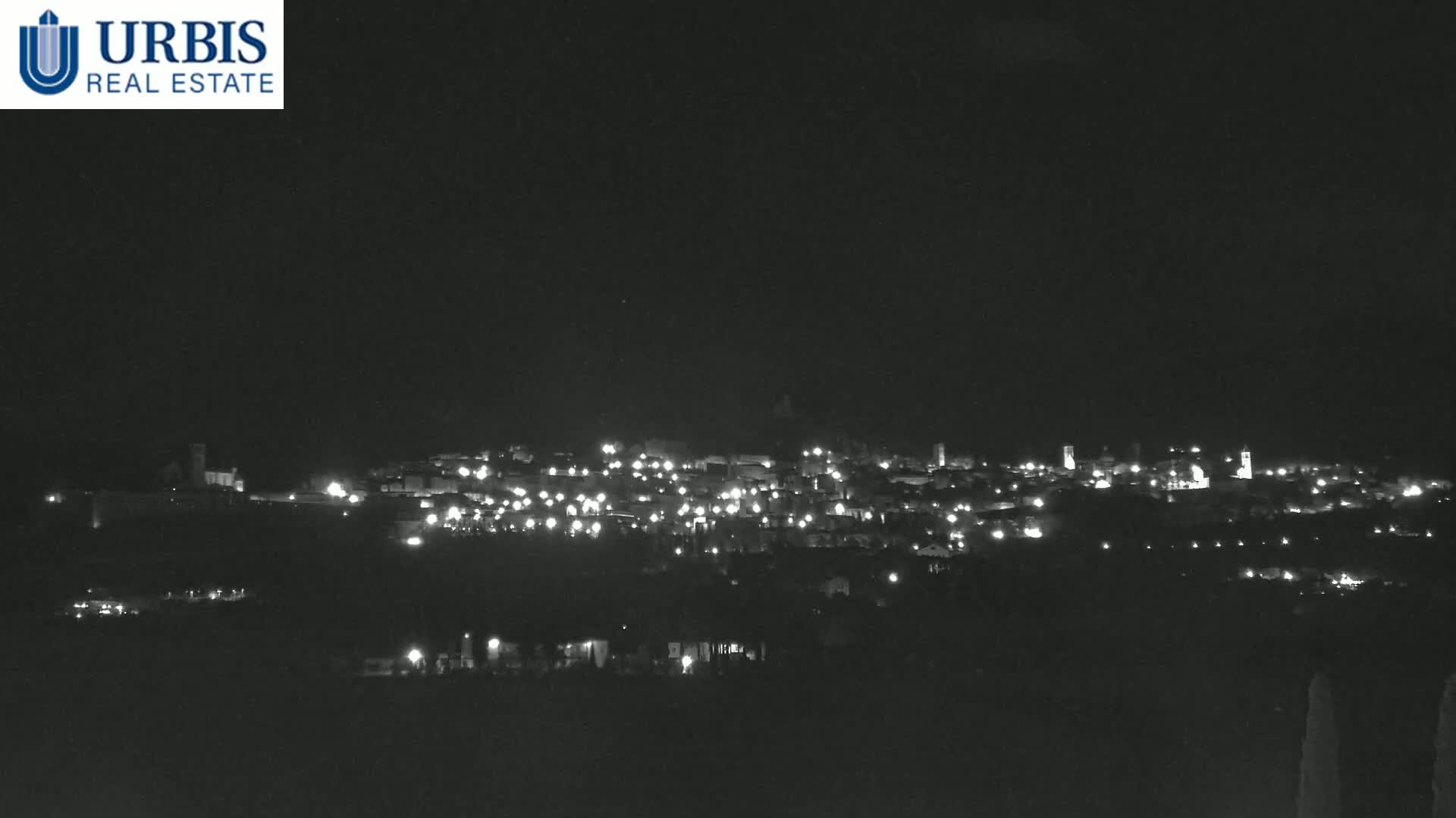 A nighttime long shot shows a small town nestled on a hill, illuminated by scattered lights under a clear, dark sky.