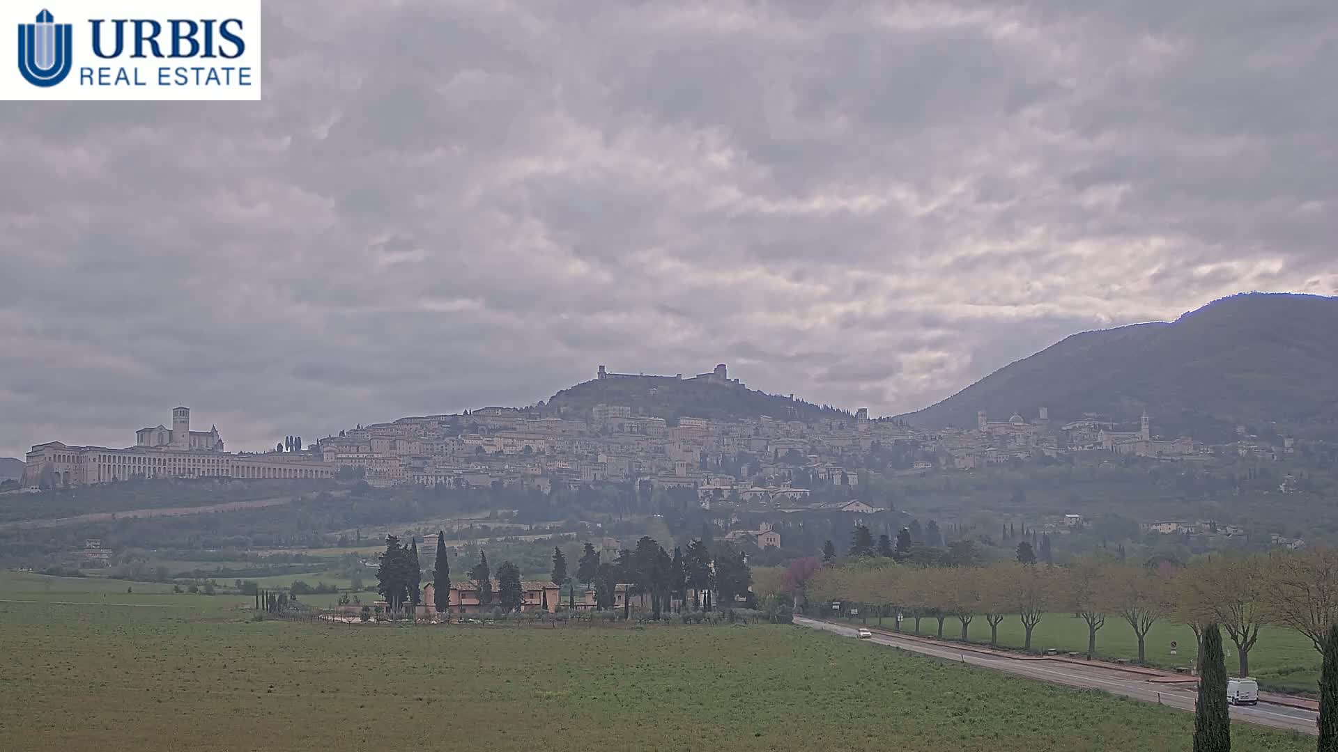 A hazy view of Assisi, Italy, nestled on a hilltop, with a long, flat building at its base, under a cloudy sky.