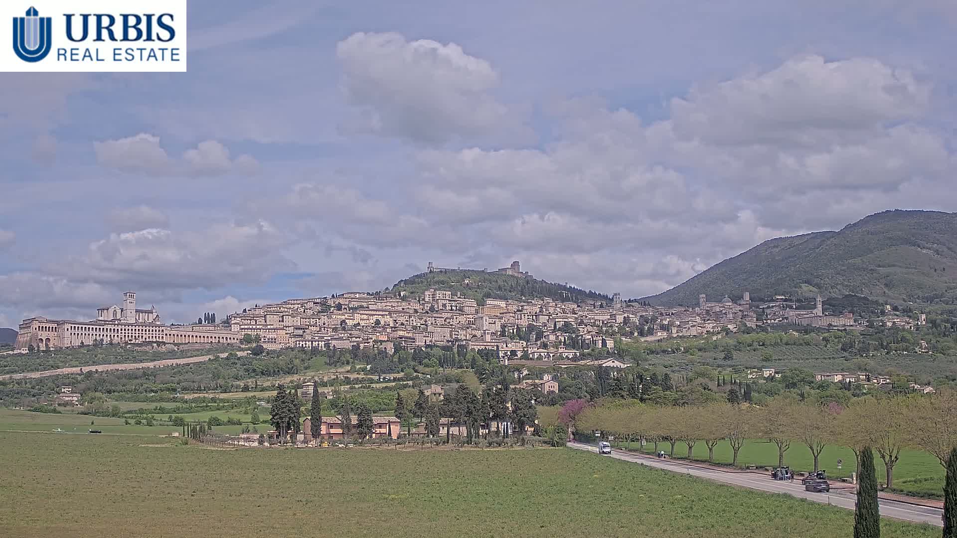 A hilltop town with a large basilica is visible under a partly cloudy sky, with a road and green fields in the foreground.