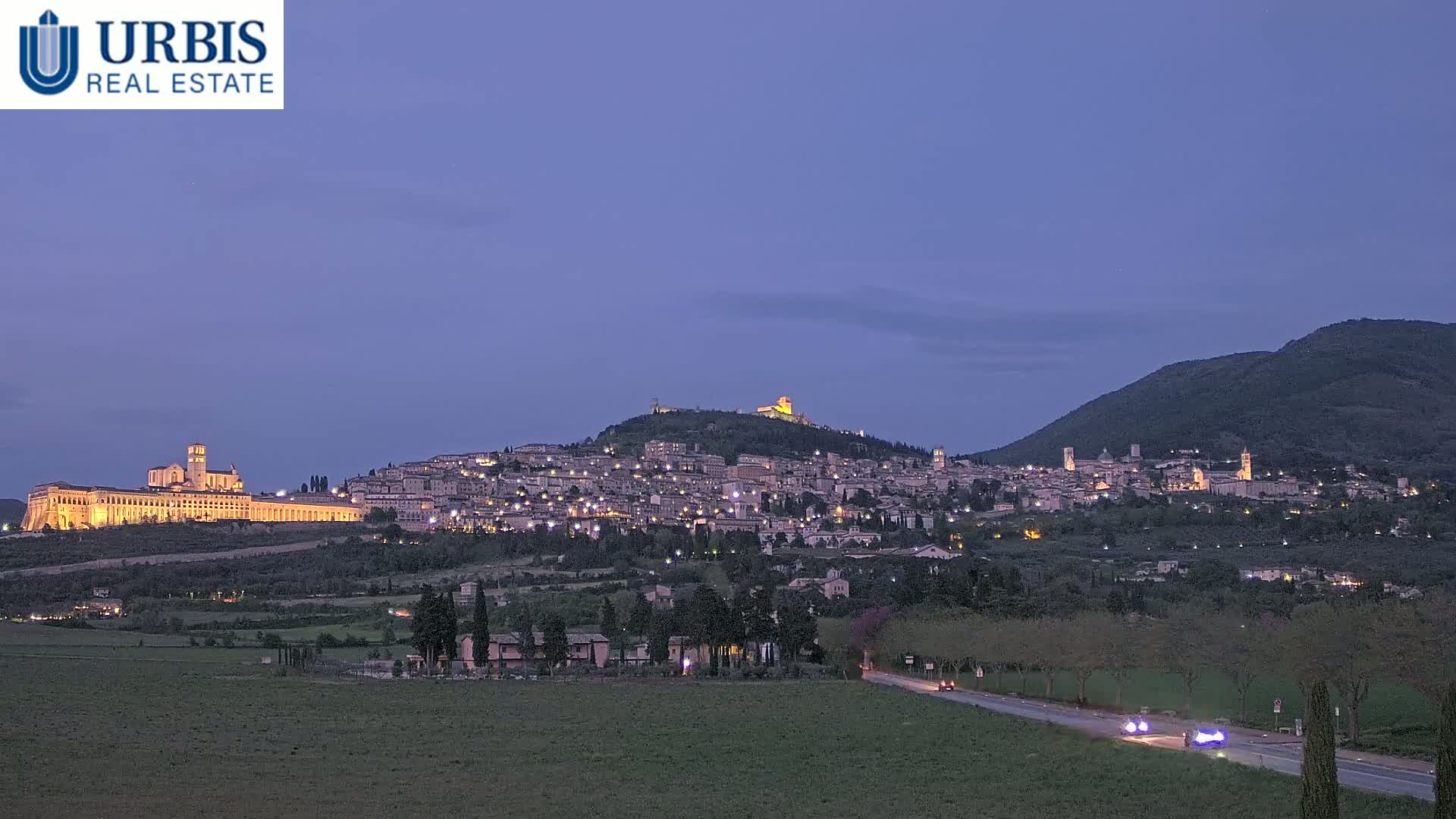 A hilltop town is illuminated at twilight, with a large basilica visible in the foreground, under a clear, dusky sky.